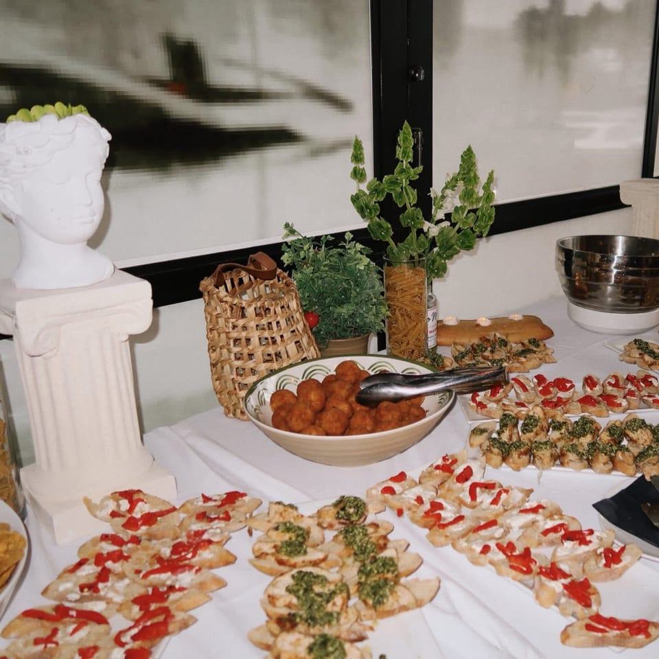 A buffet table with meatballs, bruschetta, and other appetizers, featuring a classical bust and plants.