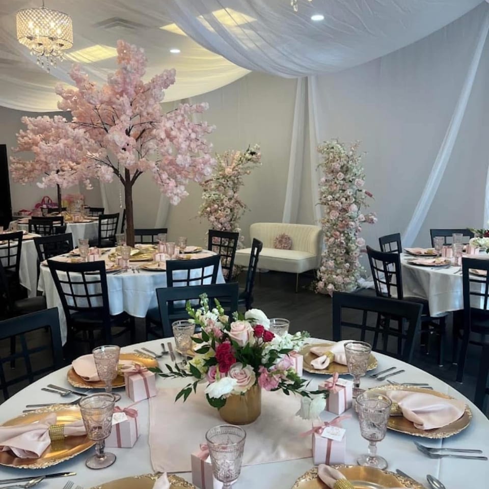 Elegant event space with round tables set with pink and gold decor, surrounded by cherry blossom trees.