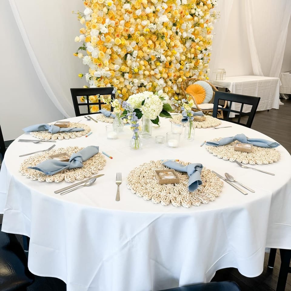A round table set for six with white tablecloths, blue napkins, and a yellow and white floral wall.