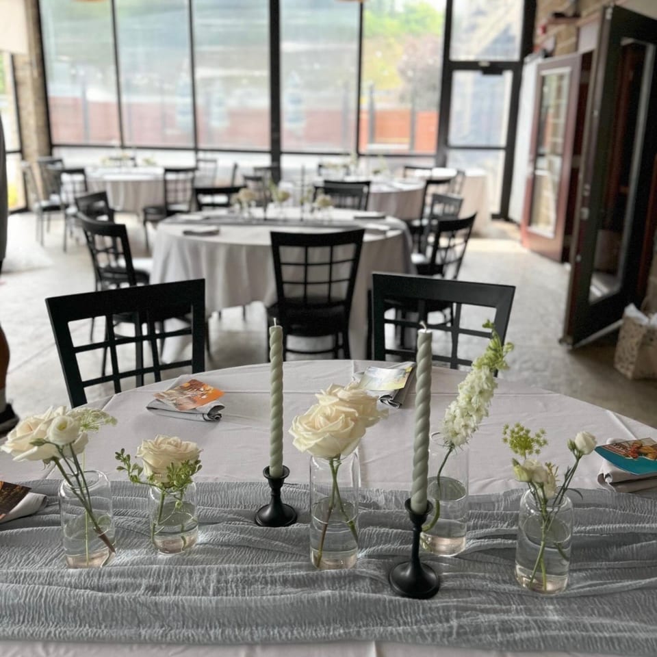Decorated table with white roses and candles, overlooking a Kavel's dining room with round tables.