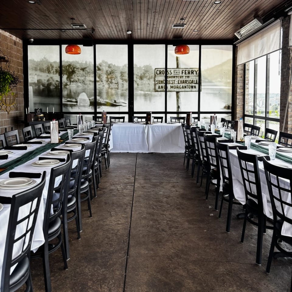 Restaurant dining room with two long tables set for a banquet, large windows overlook a river scene.