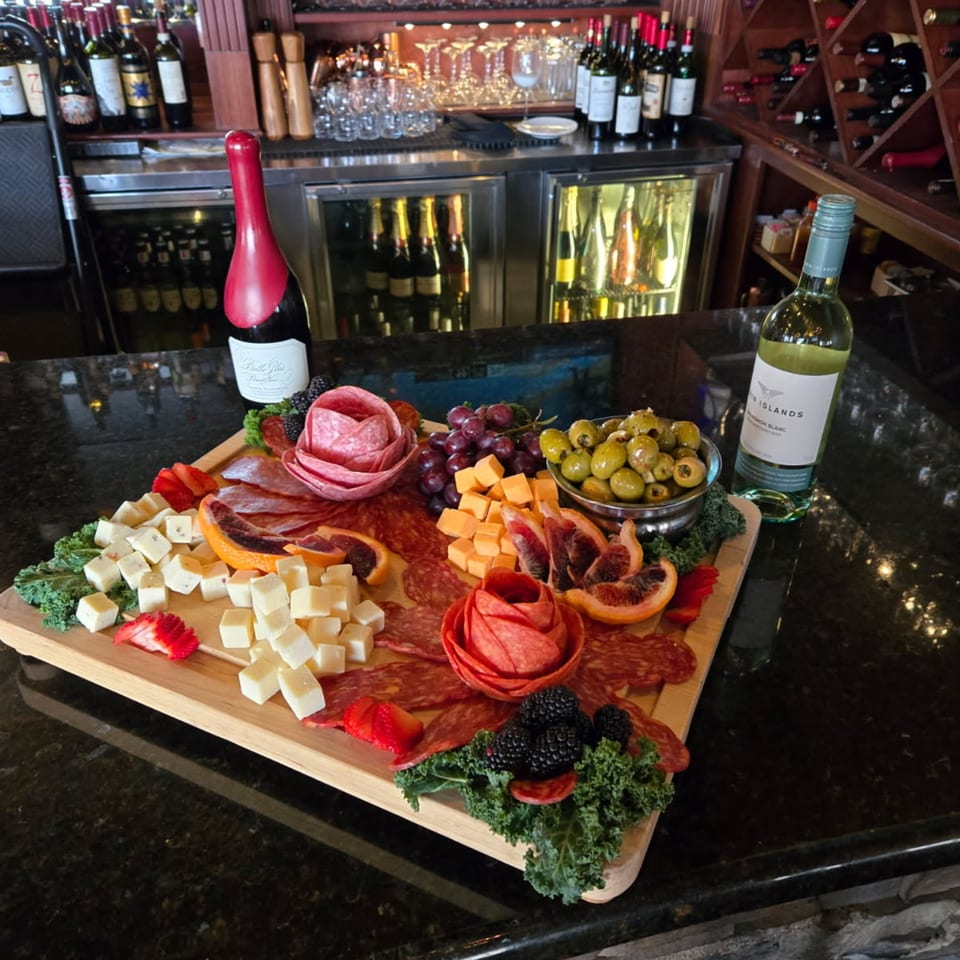 A charcuterie board with meats, cheeses, fruit, and olives sits on a bar with wine bottles.
