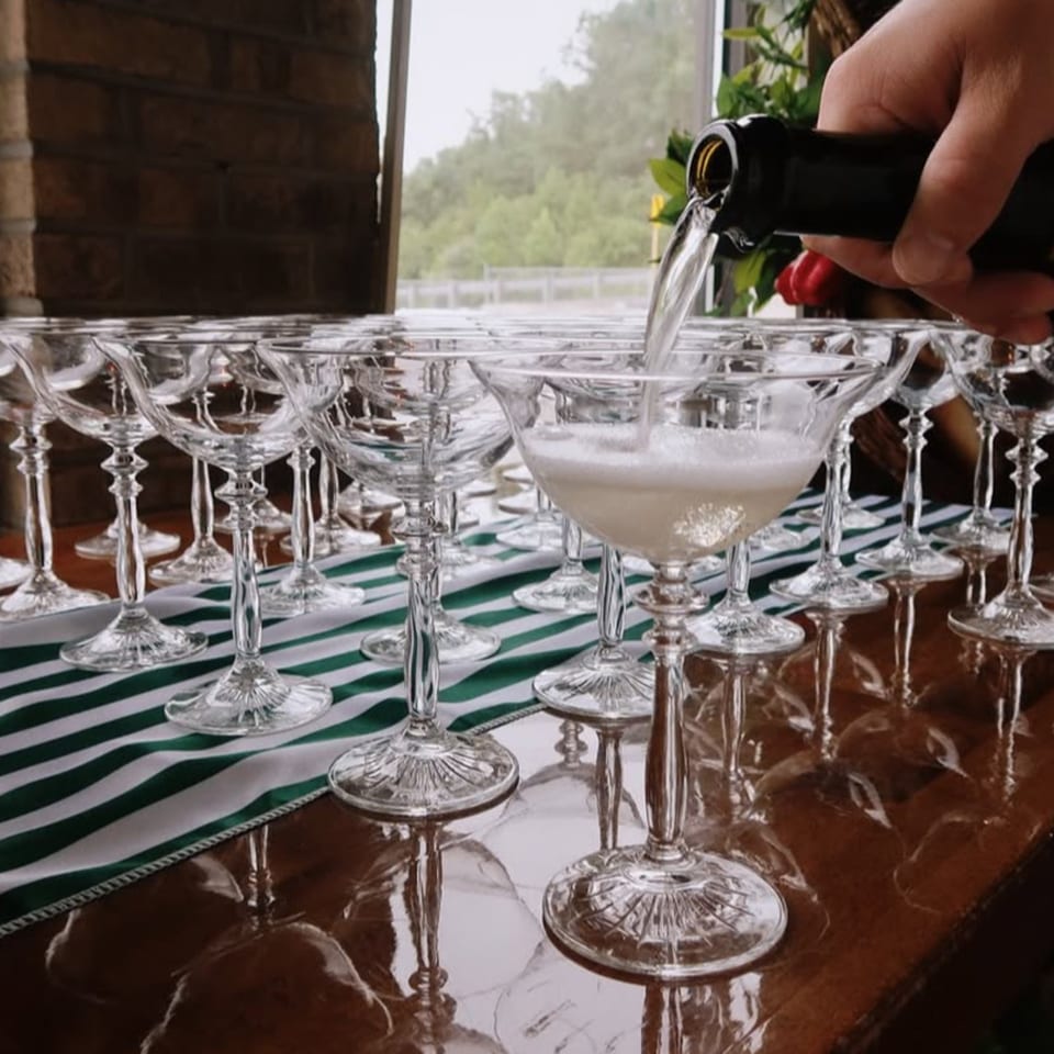 A hand pours sparkling wine into a crystal coupe glass on a table with many empty glasses.