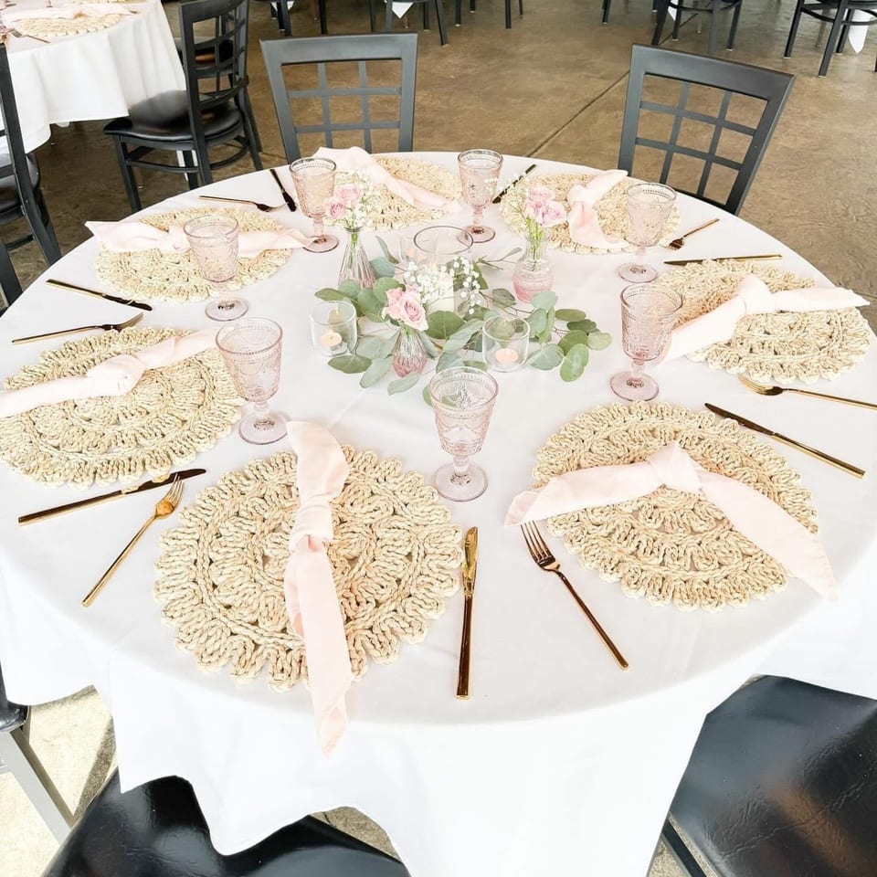 Round table set with white tablecloth, woven placemats, pink napkins, gold cutlery, and floral centerpieces.