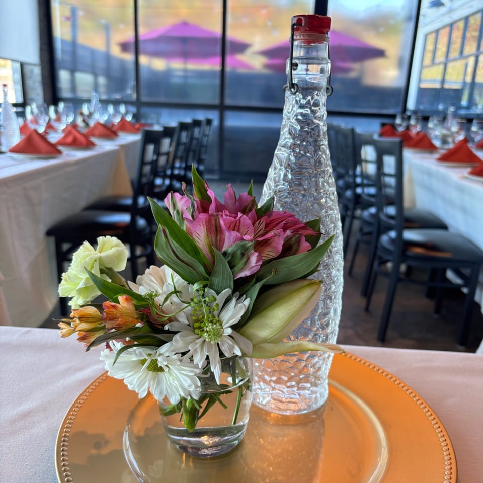 A floral centerpiece and textured glass bottle on a gold charger, with set tables in the background.