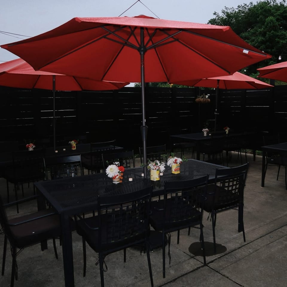 Outdoor patio with black metal tables and chairs under large red umbrellas, decorated with small flower arrangements.
