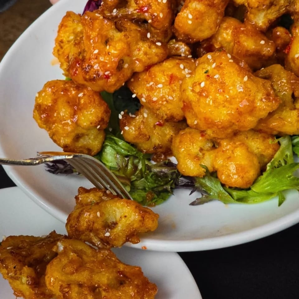 Crispy orange cauliflower bites with sesame seeds and greens on a white plate, with a fork.